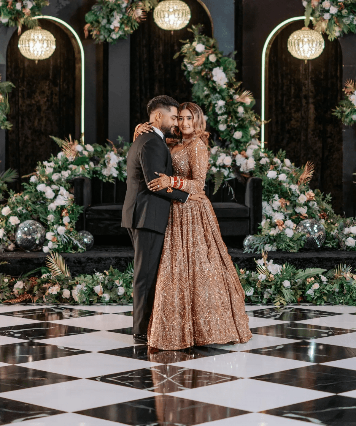 Couple dancing on elegant black and white checkered dance floor wrap with crystal chandeliers and lush floral backdrop at luxury wedding reception